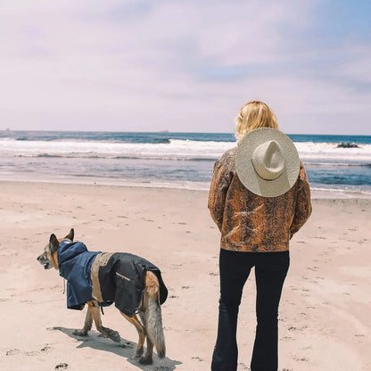 Perro con poncho impermeable Baja Coastline en la playa junto a su dueño, mirando al océano y listo para cualquier aventura costera.
