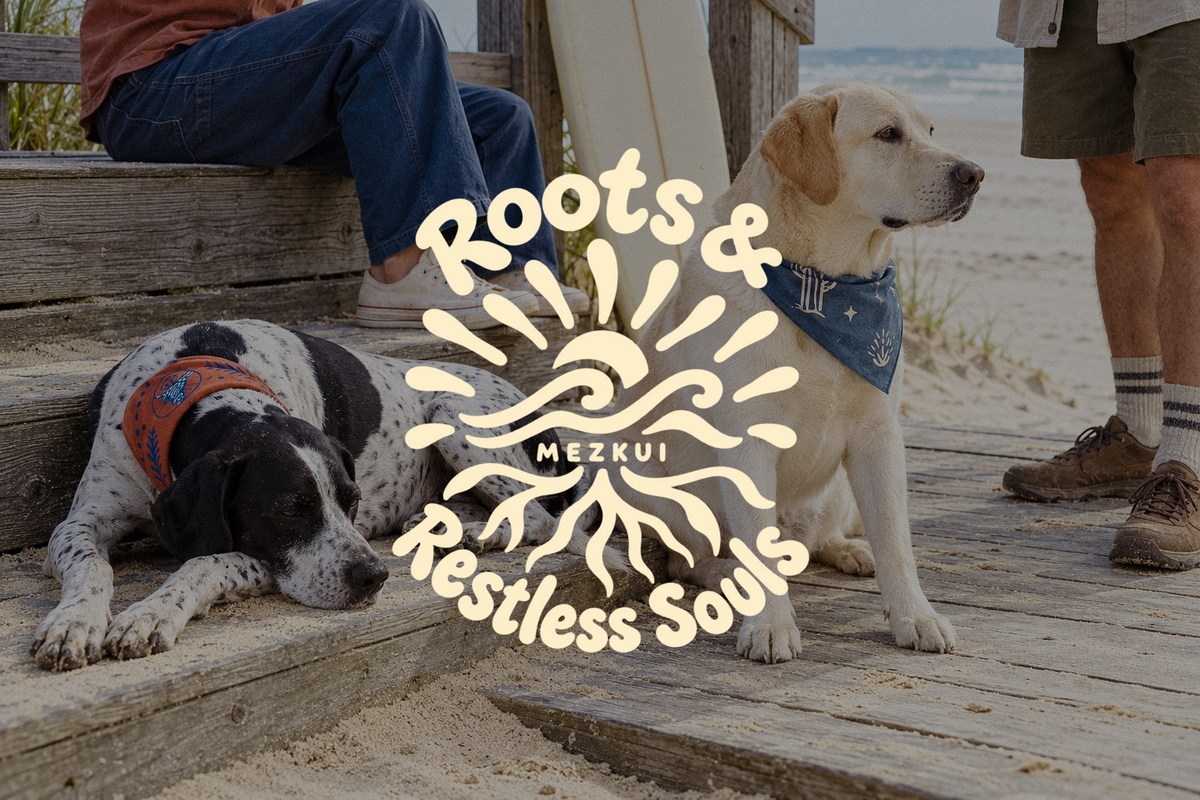 Two dogs wearing bandanas on a seaside boardwalk with people and a surfboard, beneath a cream Roots & Restless Souls MEZKUI emblem.